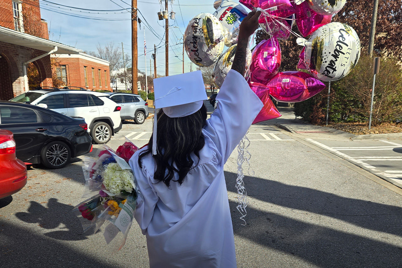 Wellspring graduate seen from behind in cap and gown, holding flowers and balloons